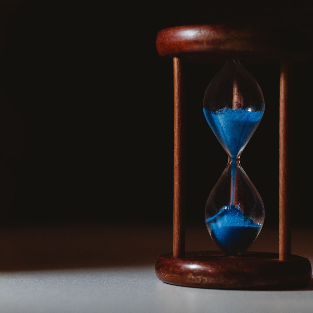 Cover art for Time Keeps Ticking: a wooden hourglass with blue sand under a spotlight